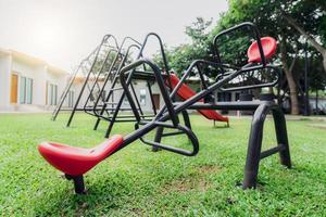 Red seesaw in the playground. Playground equipment for children to play. Plastic seesaw or teeter-totter, swing, and slide at outdoor playground with green grass ground. Outdoor kids toy at park. photo