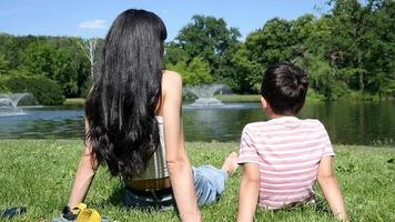 madre e hijo sentados juntos pacíficamente en la hierba verde, familia cerca del lago, día de verano video