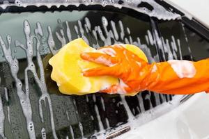 mano de mujer con guantes naranjas con esponja amarilla lavando espejo lateral coche moderno o limpiando automóvil. concepto de lavado de autos foto