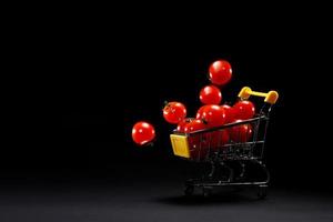 Mini shopping cart with fresh cherry tomatoes on black background. Supermarket advertisement copy space. Grocery shop. Tomatoes delivery. Transporting goods. Retail and selling. photo