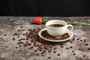 A close-up of hot coffee in a white cup is placed on a cement floor table, lots of roasted coffee beans are in the coffee cup saucer, and around, smoke and aroma waft from the cup. Blurred background
