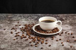 A close-up of hot coffee in a white cup is placed on a cement floor table, lots of roasted coffee beans are in the coffee cup saucer, and around, smoke and aroma waft from the cup. Blurred background