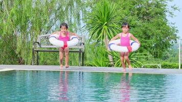 hermanitas felices con anillo de goma están saltando a la piscina al aire libre en un complejo tropical durante las vacaciones familiares de verano. niños aprendiendo a nadar. actividades saludables de verano para niños. video