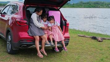 madre asiatica che guarda un video con i suoi figli su uno smartphone seduti insieme nel retro di un'auto in riva al lago. la famiglia felice, la madre e la figlia piccola trascorrono momenti felici insieme nel parco.