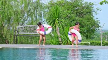 Happy little sisters with rubber ring are jumping into outdoor swimming pool in a tropical resort during family summer vacation. Kids learning to swim. Healthy Summer Activities for Kids.