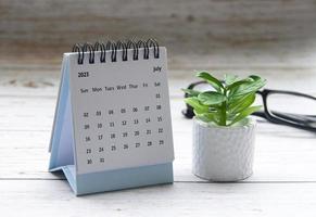 July 2023 white desk calendar on wooden table with plant and glasses background. photo