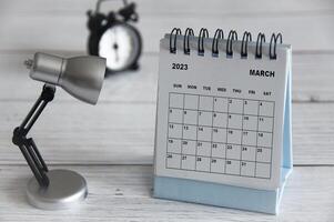 marzo de 2023 calendario de escritorio blanco con lámpara de mesa sobre mesa de madera. foto