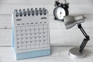 April 2023 white desk calendar with table lamp on wooden table. photo