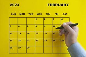 calendario de febrero de 2023 con mano sosteniendo un bolígrafo negro sobre fondo amarillo. foto