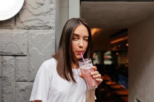 Incredible stylish woman with dark hair wearing white t-shirt is posing at camera with smoothie in city with summer drink photo