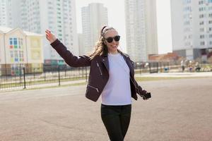 Lovely appealing girl in sunglasses wearing white t-shirt and jacket is dancing and having fun outdoor on city background photo