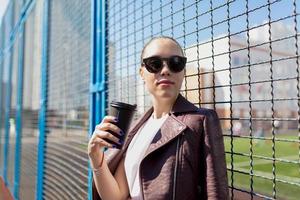 adorable dama de cabello oscuro con gafas de sol posando con café al aire libre foto