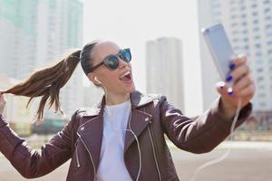 Una chica encantadora y emocionada con el pelo recogido con gafas de sol y una chaqueta de cuero está haciendo selfie en un smartphone con verdaderas emociones felices foto