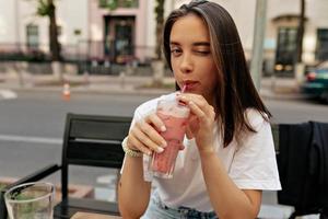 Alluring young woman winking at camera and smiling while drinking summer smoothie and sitting on terrace close-up. The bare and thin curves of collarbones are striking.
