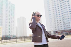 Stylish portrait of modern woman in sunglasses outdoor. Young smiling woman wearing a rock black style having fun in city. Street fashion. Sunny day. Student female going to the study photo