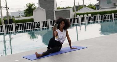 A female dressed for exercise sits on a yoga mat talking smart phone video call.