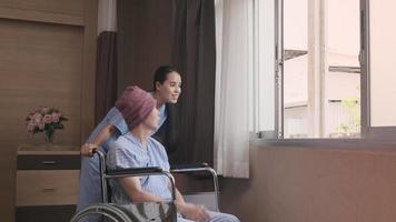 médica de terapia feminina asiática jovem uniformizada, incentivando o paciente do sexo masculino em cadeira de rodas na janela para apoiar e motivar a recuperação, doença do câncer após tratamento médico de quimioterapia na sala de internação do hospital. video