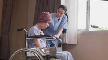 Una joven y uniformada médica de terapia asiática con un paciente varón en silla de ruedas en la ventana para apoyar la recuperación, sufrir y toser de la enfermedad del cáncer después del tratamiento médico con quimioterapia en la habitación del hospital. video