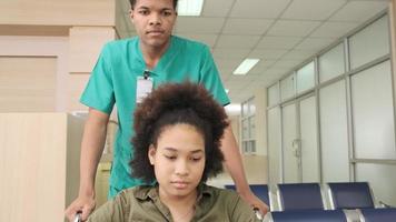 Young African American female doctor with stethoscope in uniform checks injury patient girl in wheelchair at outpatient accident clinic hospital, illness medical clinic examination, healthcare hall. video