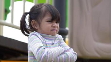 Handheld shot, Portrait Asian girl sits frowning arms crossed with angry on slider in playground video