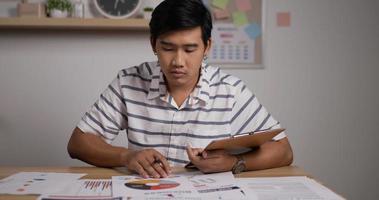 Portrait of Asian bussinesman holds pen makes notes on sheet of paper signs document. Young male working studying from home sitting at table.