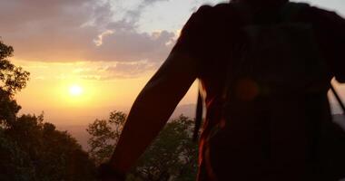 tiro retroiluminado en cámara lenta, vista trasera de cerca joven excursionista caminando, levantando las manos y mirando alrededor con felicidad en la cima de la montaña rocosa al atardecer video