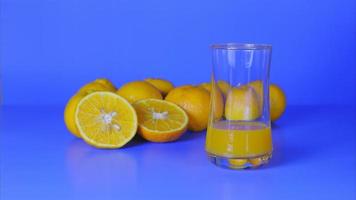 Stop motion vivid color studio shot, orange juice level in glass increasing, and group of natural fresh oranges on blue background, organic beverage, healthy refreshment, and tasty nutrient drink.