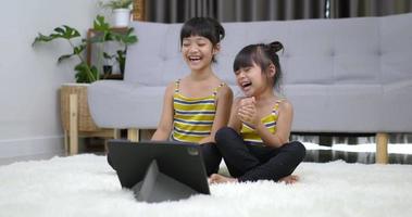 toma en cámara lenta, dos encantadoras chicas asiáticas disfrutan y se ríen mientras ven una película en una tableta en la sala de estar