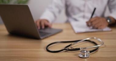 Close up of A stethoscope is placed on table with doctor working on laptop and making note background. Medical equipment concept video