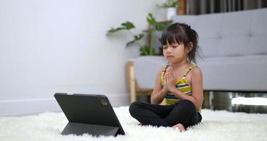 tiro em câmera lenta, menina asiática sentada no tapete fechou o olho e colocou as duas mãos no peito em meditação asana pose treinamento no tablet na sala de estar em casa