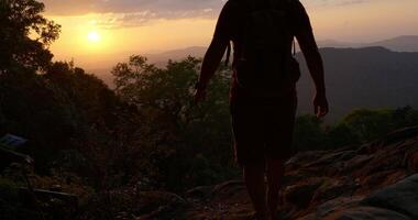 colpo retroilluminato, vista posteriore giovane escursionista che cammina e alza le mani con felice sulla cima della montagna rocciosa nel tramonto video