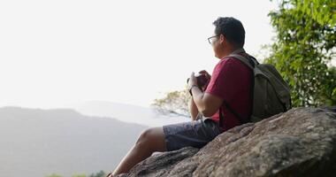 colpo al rallentatore, giovane viaggiatore con zaino e sacco a pelo in occhiali usa la fotocamera digitale per scattare una foto mentre era seduto sulle scogliere rocciose nella foresta video