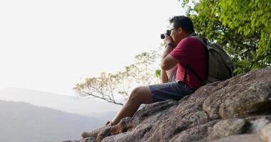 Zeitlupenaufnahme, junger Rucksacktourist mit Brille, der eine Digitalkamera verwendet, um ein Foto zu machen, während er auf den felsigen Klippen im Wald sitzt video