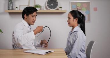 Portrait of young Asian doctor hold stethoscope exam female patient in clinic office. Male doctor check heart lungs of woman checkup in hospital. Medical and health care concept. video