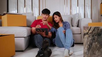 Slow motion shot, Young couple sitting front of sofa on floor and use tablet while counting boxs after relocation in their new home, new house concept video