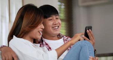 Slow motion shot, Young couple sitting on sofa in living room and use smartphone playing game together, they talk and laughing with happiness, happy family concept video