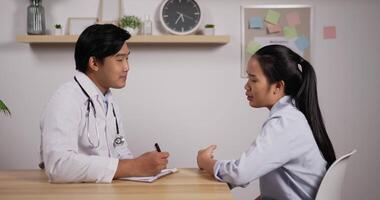 retrato de un joven médico asiático y un paciente sentados y discutiendo los resultados del examen de salud en el consultorio de la clínica. encantador médico masculino dando consejos a una paciente. concepto de atención médica y de salud. video