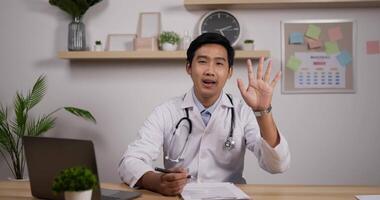 Portrait of young asian male doctor with stethoscope making online video call consult patient and waving hand. Medical assistant therapist videoconferencing. Telemedicine pandemic concept.