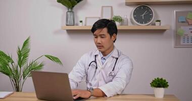 porträtt av ung asiatisk manlig läkare kardiolog bär vit medicinsk rock att skriva laptop och visar inga tecken på klinikkontoret. medicinsk och hälsovård koncept. video