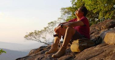 slow-motion shot, jonge backpacker man houdt digitale camera vast en kijkt rond terwijl hij op de rotsachtige kliffen in het bos zit met zonlicht video