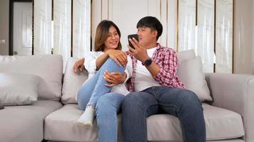 tir au ralenti, heureux jeune couple assis sur un canapé dans le salon à la maison utiliser un smartphone discuter avec un ami, parler et rire avec bonheur, concept de famille heureuse