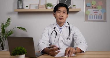 retrato de jovem médico masculino asiático com estetoscópio faz videochamada online consultar paciente e olha para a câmera. videoconferência do terapeuta assistente médico. conceito de pandemia de telemedicina.