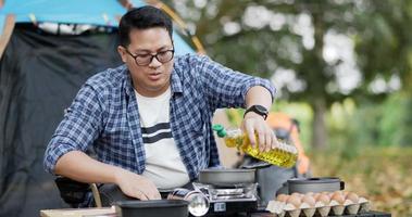 retrato de un viajero tailandés con gafas vertiendo aceite de girasol en una sartén. cocina al aire libre, viajes, camping, concepto de estilo de vida. video