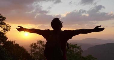 Hintergrundbeleuchtung in Zeitlupe, Nahaufnahme von hinten junger Wanderer steht, Hände hoch und schaut sich glücklich auf dem Gipfel des felsigen Berges im Sonnenuntergang um