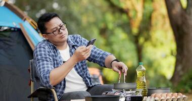 portret thai reiziger man bril met smartphone tijdens het koken van een maaltijd op een camping. buiten koken, reizen, kamperen, lifestyle concept. video