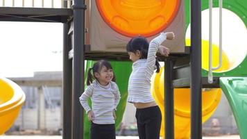 Two Asian little children standing and raising arms over head, chatting and laughing together happily at playground