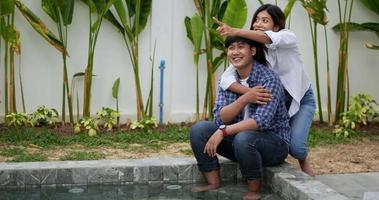 une femme asiatique embrasse son mari par derrière, se pointe joyeusement du doigt pour regarder la nature au bord de la piscine, parle et rit avec bonheur. notion de famille heureuse