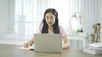 jeune femme asiatique école étudiante porter des écouteurs apprendre à regarder la classe de webinaire en ligne en regardant le cours à distance d'apprentissage en ligne sur ordinateur portable ou l'enseignant d'appel vidéo par webcam à la maison. video