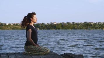 vista laterale di una giovane donna snella seduta su un molo in posizione di loto e alzando le mani. donna che pratica yoga sulla spiaggia al tramonto contro l'acqua. Video al rallentatore 4K.