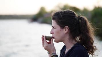 une jeune femme boit du thé au coucher du soleil sur le rivage. Vidéo au ralenti 4k. video